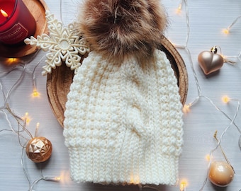 Puede incluir: Gorro de invierno de punto crema, tejido a mano, con diseño de trenza y un gran pompón marrón esponjoso. El gorro se muestra sobre un plato de madera, rodeado de luces decorativas y adornos, creando una escena festiva.