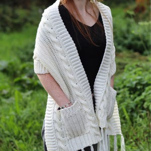 Puede incluir: Chal de punto de color crema con dos bolsillos delanteros y flecos. El chal está hecho de un hilo suave y texturizado y presenta un diseño clásico de punto de cable. Un accesorio acogedor y elegante.