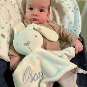 Baby holding his personalised white bunny baby comforter with baby name embroidered in light blue. Underside is a light grey colour. Square shaped with a bunny head attached in the center. Corners are knot tied. Comforter has a gentle rattle.