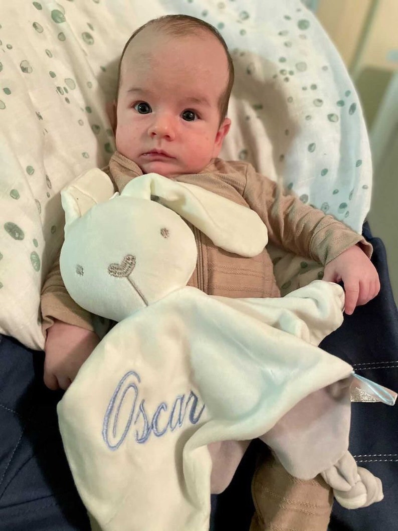 Baby holding his personalised white bunny baby comforter with baby name embroidered in light blue. Underside is a light grey colour. Square shaped with a bunny head attached in the center. Corners are knot tied. Comforter has a gentle rattle.