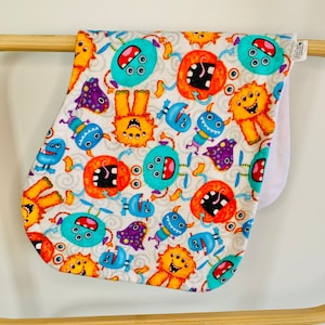 Puede incluir: Un babero blanco para bebé con un patrón colorido de monstruos de dibujos animados con múltiples ojos, bocas y piernas. Los monstruos son en tonos de naranja, azul, morado y amarillo.