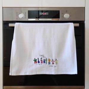 May include: White kitchen towel featuring embroidered figures of people in different colours. The text "Rogers Family" and "by meid 2013" are embroidered on the towel. The towel is hanging on an oven door.
