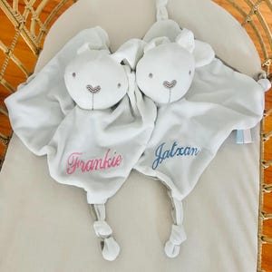 Two white bunny baby comforters in a cot. One personalized with Frankie in light pink thread and the other embroidered with Jatxan in light blue thread.