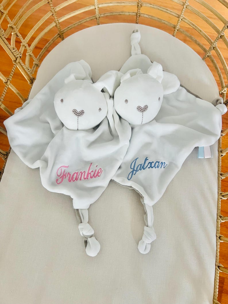 Two white bunny baby comforters in a cot. One personalized with Frankie in light pink thread and the other embroidered with Jatxan in light blue thread.
