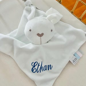 A personalised baby comforter embroidered with Ethan in blue thread. The comforter is a white bunny comforter placed in a baby cot.