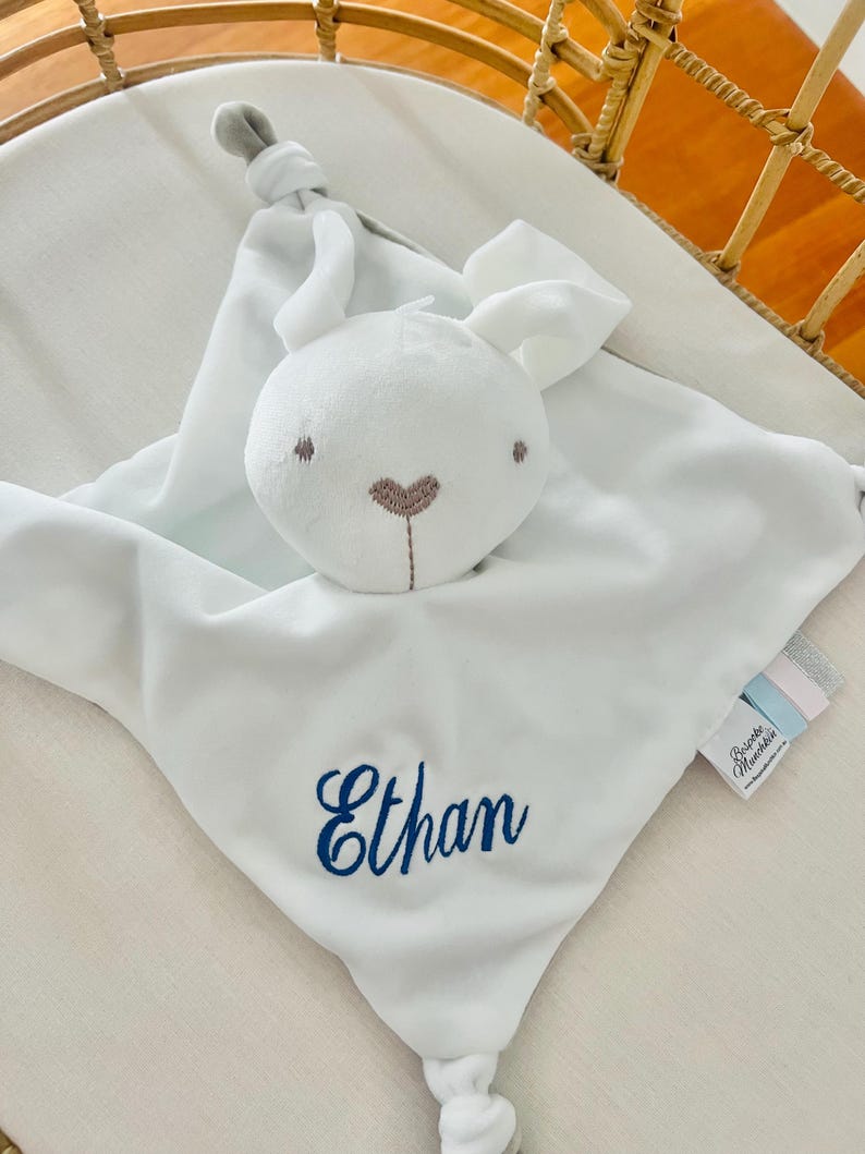A personalised baby comforter embroidered with Ethan in blue thread. The comforter is a white bunny comforter placed in a baby cot.