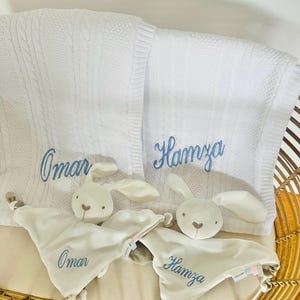 Two sets of personalised baby gifts consisting of a baby blanket and a white bunny baby comforter. One set with Omar embroidered in light blue thread and the other set with Hamza, also in light blue thread. The baby blankets are draped over a cot with the baby comforters resting in the cot against the baby blankets.