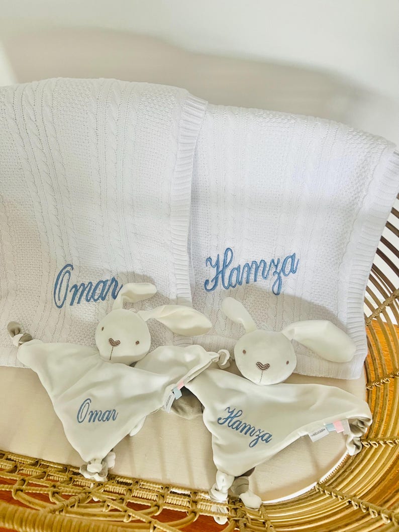 Two sets of personalised baby gifts consisting of a baby blanket and a white bunny baby comforter. One set with Omar embroidered in light blue thread and the other set with Hamza, also in light blue thread. The baby blankets are draped over a cot with the baby comforters resting in the cot against the baby blankets.