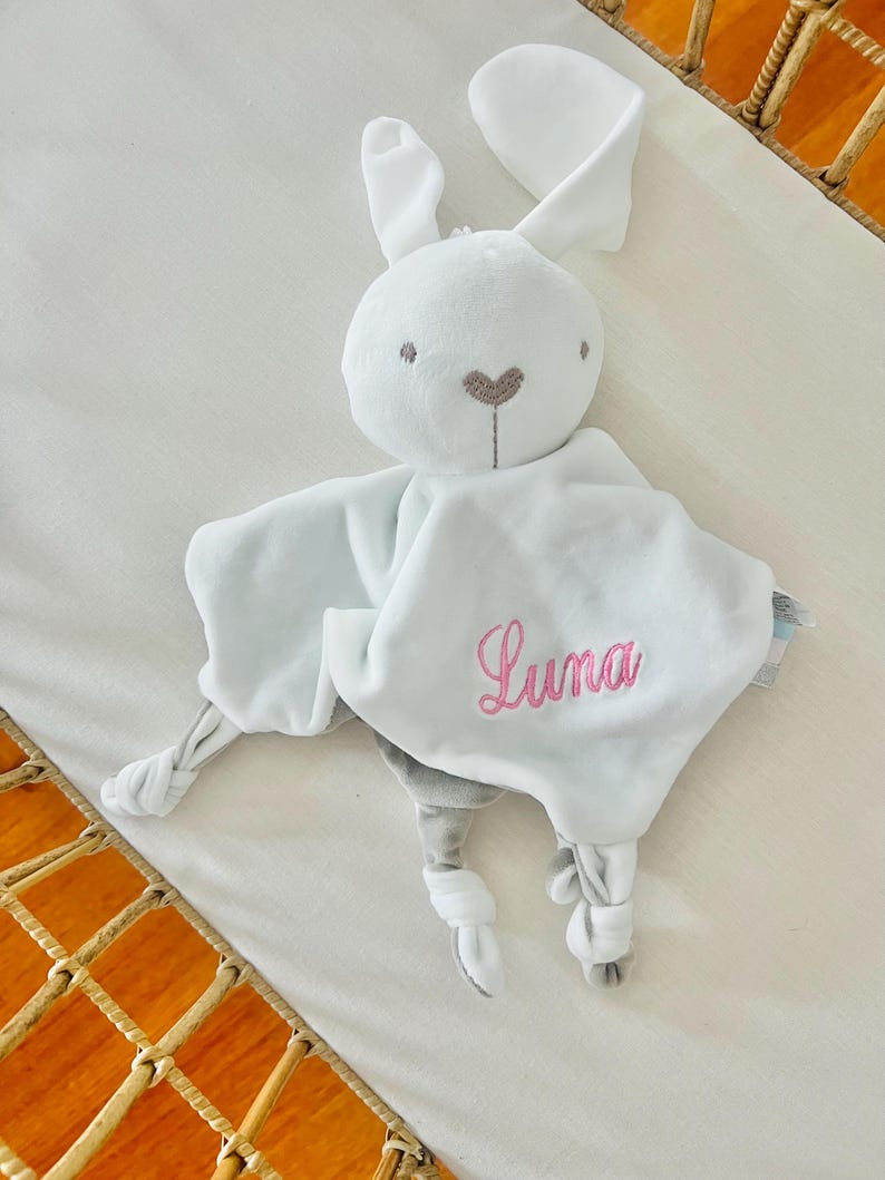 Personalised white bunny baby comforter embroidered with Luna in light pink thread. Baby comforter in is a cot.