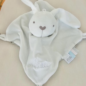 White personalised baby comforter security blanket embroidered with Maxine in white. Baby comforter is in a cot.