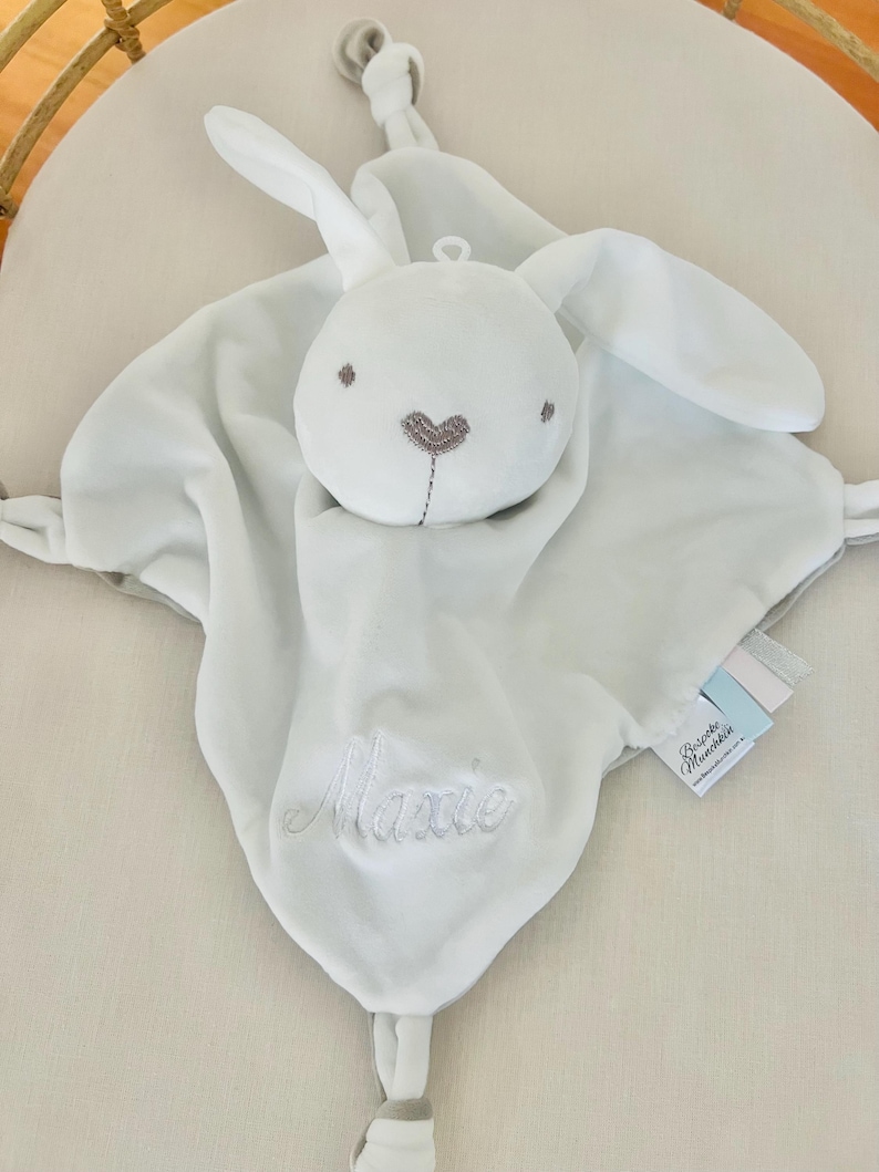 White personalised baby comforter security blanket embroidered with Maxine in white. Baby comforter is in a cot.