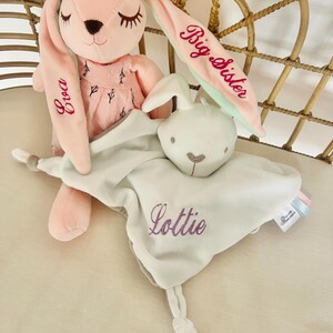 White personalised bunny baby comforter personalised with name Lottie in purple, resting on a pink bunny with Eva embroidered one one ear and Big Sister on the second ear. Both plush bunny and baby comforter are in a cot.