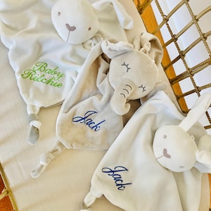 Three personalised baby comforters embroidered with baby name lined up in a cot. A white bunny comforter with Baby Ritchie in green, elephant baby comforter with Jack in blue and a white bunny comforter also with Jack in blue.