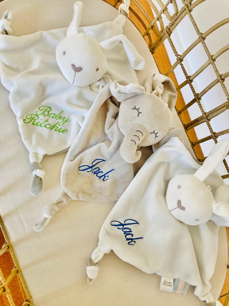 Three personalised baby comforters embroidered with baby name lined up in a cot. A white bunny comforter with Baby Ritchie in green, elephant baby comforter with Jack in blue and a white bunny comforter also with Jack in blue.
