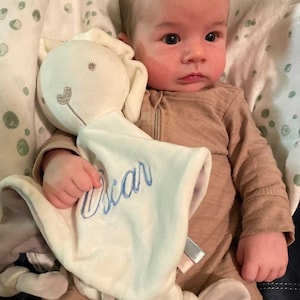 Baby holding his personalised white bunny baby comforter with baby name embroidered in light blue. Underside is a light grey colour. Square shaped with a bunny head attached in the center. Corners are knot tied. Comforter has a gentle rattle.
