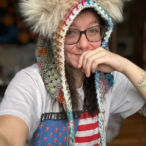 May include: A colorful crocheted hooded hat with faux fur earmuffs. The hat has a long, braided tassel and is made with a variety of colors, including pink, blue, green, and white.