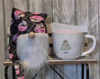 Puede incluir: Un gnomo caprichoso con una larga barba blanca y una nariz de madera está sentado junto a una taza blanca. El sombrero del gnomo tiene un estampado de tazas de café rosas y personajes de malvavisco. La taza presenta un diseño de pastel y la palabra "LOVE."
