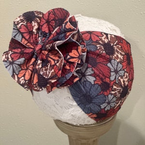 May include: A floral headband featuring a large, ruffled flower in shades of red, brown, and blue. The headband is made from a soft, stretchy fabric and is ideal for adding a touch of whimsy to any outfit.