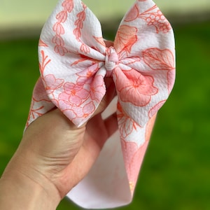 May include: A white headband with a pink floral print. The headband is tied in a bow and is made of a soft, textured fabric.