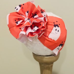May include: A coral pink and white headband with a large, ruffled bow. The headband has a ghost pattern with black eyes and mouths.