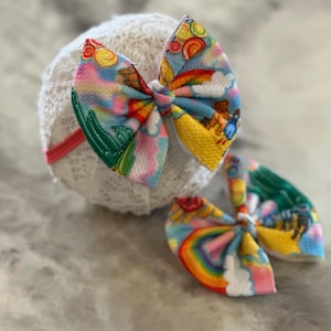 May include: Two colorful hair bows with a rainbow and yellow brick road design. The bows are made of fabric and have a white background with a rainbow, clouds, and a yellow brick road.