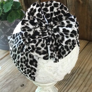 May include: A white headband with a black and white leopard print bow. The bow is made of fabric and has a ruffled edge.