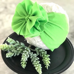 May include: A lime green knotted headband with a large, ruffled bow. The headband is on a white lace background.