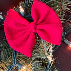 May include: A bright red bow made of a textured fabric. The bow is sitting on a green artificial Christmas tree branch.