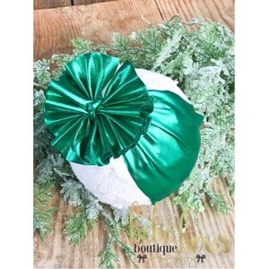 May include: A green metallic bow with a white background. The bow is made of fabric and has a ruffled texture.