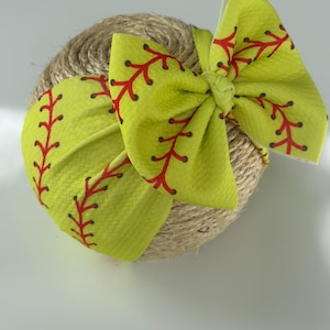 May include: A bright yellow headband with a softball design. The headband features a bow and is decorated with red stitching, mimicking a softball. The headband is wrapped around a ball of twine.