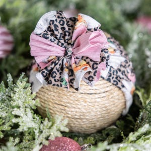 May include: A baby headband with a large bow. The bow features a mix of patterns, including leopard print, floral, and solid pink fabric. The headband is wrapped around a ball of twine, with greenery and pink baubles in the background.