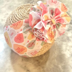 Peut inclure: Un bandeau pour bébé avec un nœud et une base enveloppée. Le bandeau présente un tissu blanc avec des graphiques de cœurs de bonbons colorés et les phrases "Dream Big", "Text Me" et "You Rock". Le nœud est rose et blanc.