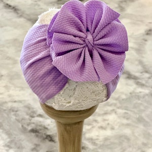 Peut inclure: Un bandeau turban lavande avec un grand nœud plissé. Le bandeau est présenté sur un support en bois.