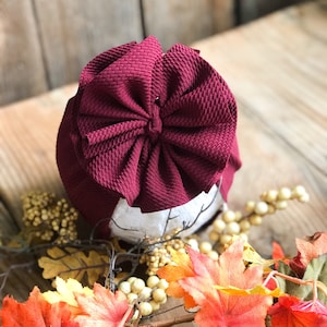 May include: A burgundy turban with a large bow on top. The turban is made of a soft, textured fabric. The turban is sitting on a wooden surface with autumn leaves.