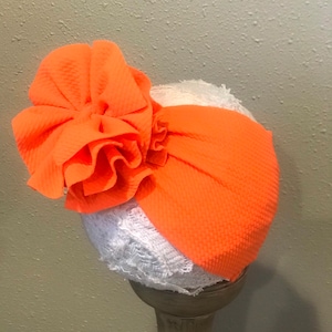Peut inclure: Un bandeau turban orange avec une grande fleur en tissu orange sur le dessus. Le bandeau est sur une tête de mannequin recouverte de dentelle blanche.