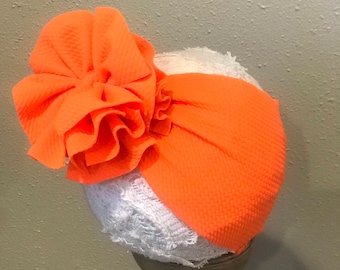 May include: Orange turban headband with a large orange fabric flower on top. The headband is on a white lace covered mannequin head.