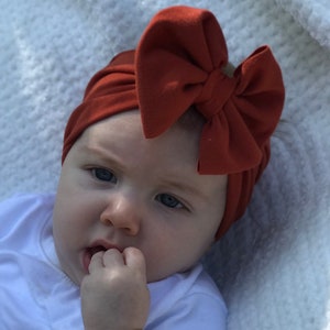 Könnte beinhalten: Ein Baby trägt einen turban in Rostfarbe mit einer großen Schleife oben.