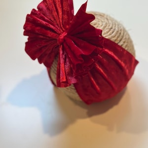 Op de afbeelding: Een felrode baby haarband van fluweel met een grote strik. De strik heeft een gerimpeld ontwerp en een zachte textuur. De haarband is gewikkeld rond een bol touw.