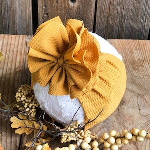 Peut inclure: Un bandeau en tissu jaune moutarde avec un grand nœud plissé. Le bandeau est fait d'un matériau doux et extensible et est parfait pour ajouter une touche de style à n'importe quelle tenue.