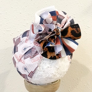 May include: A white fabric headband with a large, ruffled bow. The bow is made of red, white, and blue fabric with a leopard print pattern. The headband is decorated with a white lace trim.