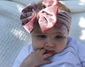 May include: A baby wearing a dusty rose velvet headband with a large bow on top. The headband is tied around the baby's head and the bow sits on top of the baby's forehead. The baby is wearing a white t-shirt and has blue eyes.