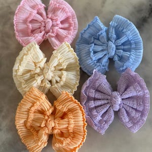 Könnte beinhalten: Sechs Haarschleifen aus Stoff in Pastellfarben: Rosa, Hellblau, Gelb, Orange und Lavendel. Die Schleifen haben ein strukturiertes, plissiertes Design und sind in der Mitte gebunden. Geeignet für Babys und Kleinkinder.