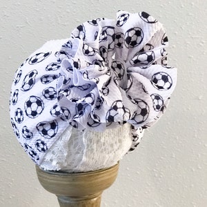 May include: White fabric headband with a large ruffle made of white fabric with a black soccer ball print. The headband is displayed on a wooden stand.