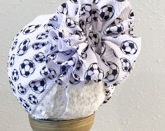 Puede incluir: Diadema de tela blanca con un gran volante de tela blanca con estampado de balón de fútbol negro. La diadema se muestra en un soporte de madera.