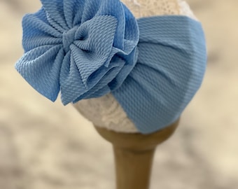 May include: A light blue turban headband with a large, fluffy bow. The headband is made of a soft, stretchy fabric and has a lace trim.