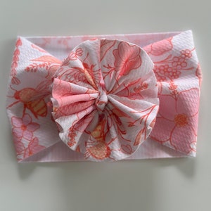 May include: A pink and white headband with a floral print and a large, knotted bow. The headband features a pattern of pink flowers, leaves, and bees.