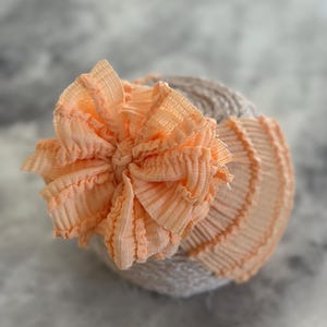 May include: A peach-coloured, ruffled fabric flower sits atop a light grey, textured headband. The flower is the central focus. The background is a soft, blurred grey.