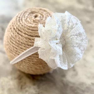 May include: A white lace headband featuring a large, ruffled flower. The headband is made of white fabric and is designed to be worn around the head. The flower is the focal point, adding a delicate and feminine touch to the accessory.