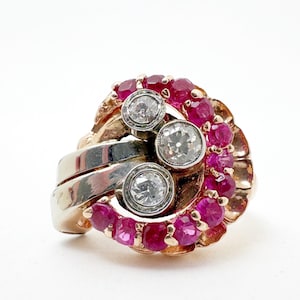 Peut inclure: Une bague en or au design incurvé, ornée de trois pierres claires rondes disposées verticalement, entourées d'un arrangement semi-circulaire de petits rubis rose vif. L'anneau présente une finition en or poli avec une structure large et lisse.