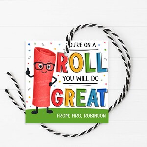 May include: A square gift tag with a red candy roll character wearing glasses and giving a thumbs up. The tag has the words "YOU'RE ON A ROLL YOU WILL DO GREAT" in colorful letters, with "FROM: MRS. ROBINSON" at the bottom. A black and white string is attached.
